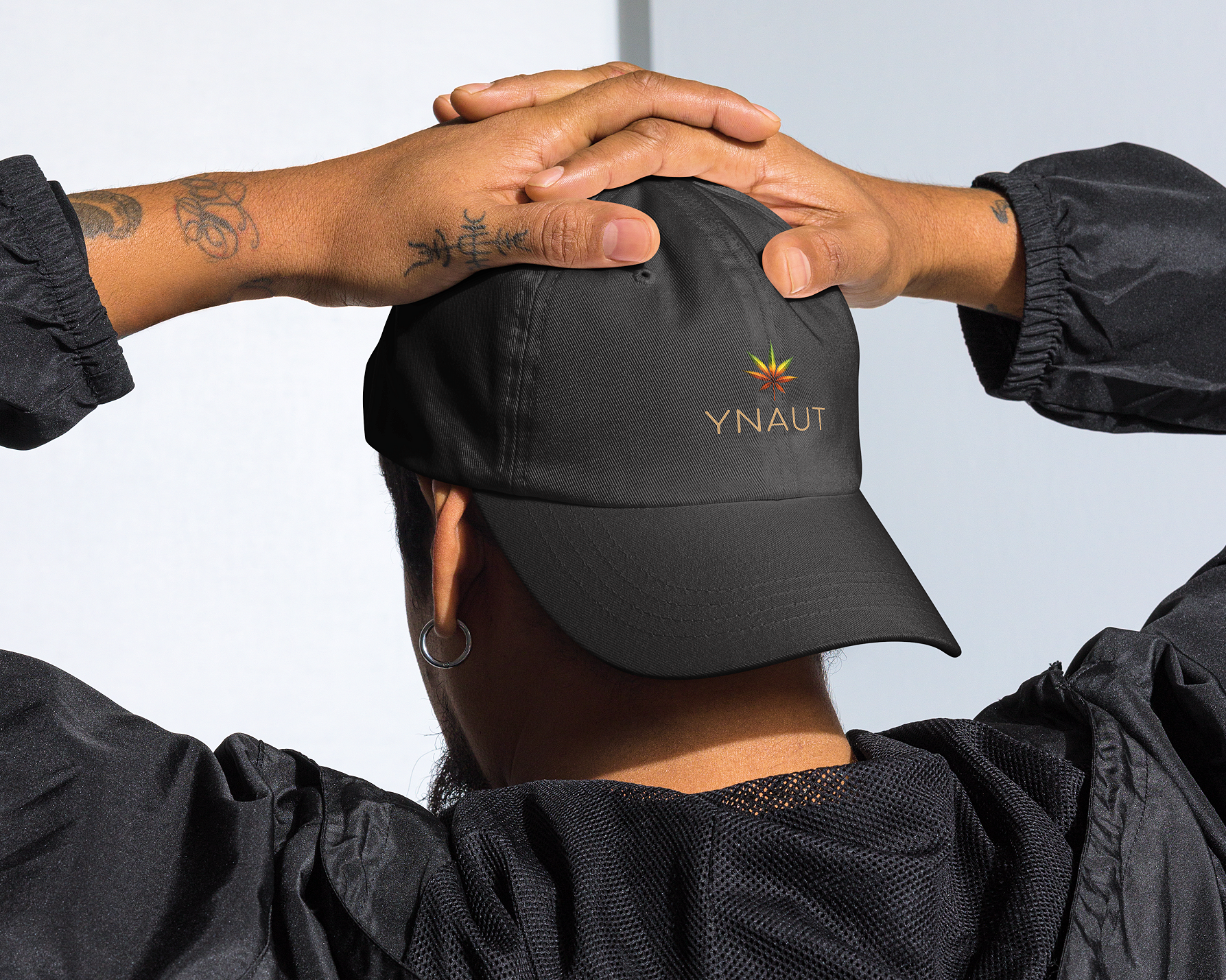 Person wearing a black cap with 'YNAUT' logo, hands clasped behind head against a light background