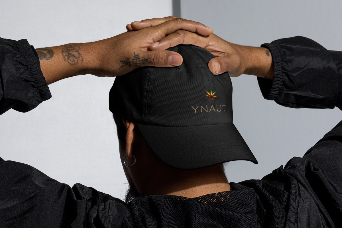 A person wearing a black baseball cap with 'YNAUT' embroidered on the front, shown from behind with their hands resting on the cap.